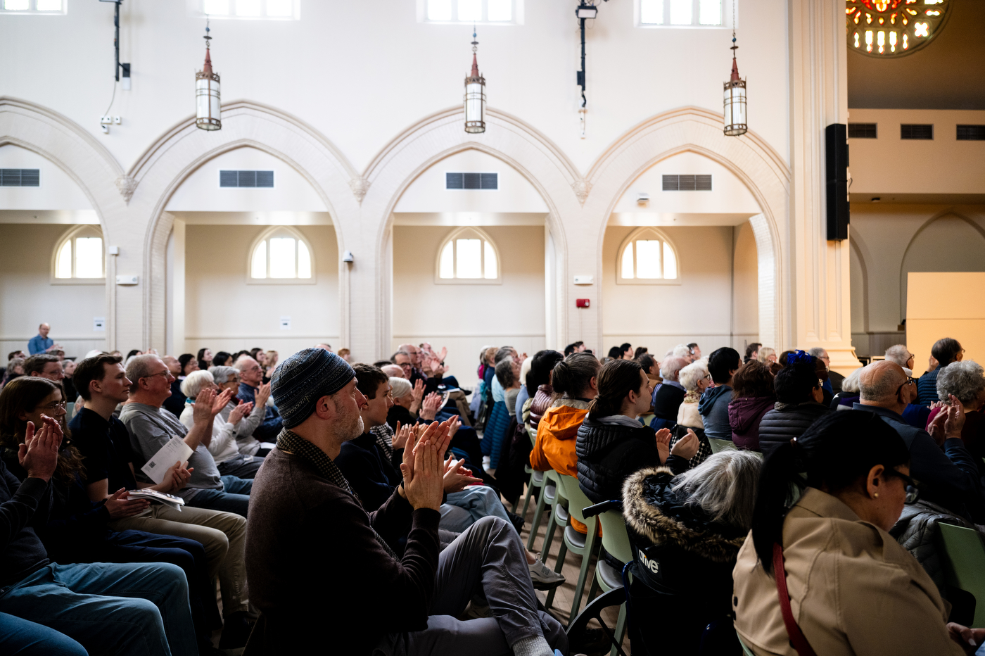 A crowd applauds in a room with vaulted cielings and stain glass windows.