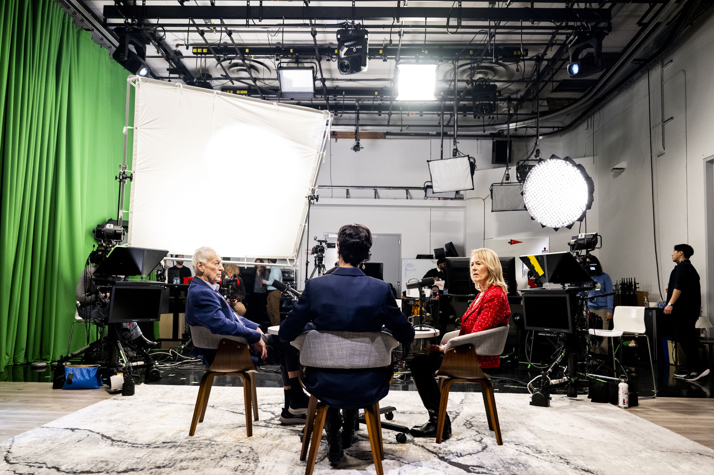 Daniel McCarthy, Ann Simmons and Sheila Puffer sit in conversation on set at the Northeastern University NGN studio, surrounded by cameras, lighting rigs and a green screen, as crew members work in the background.