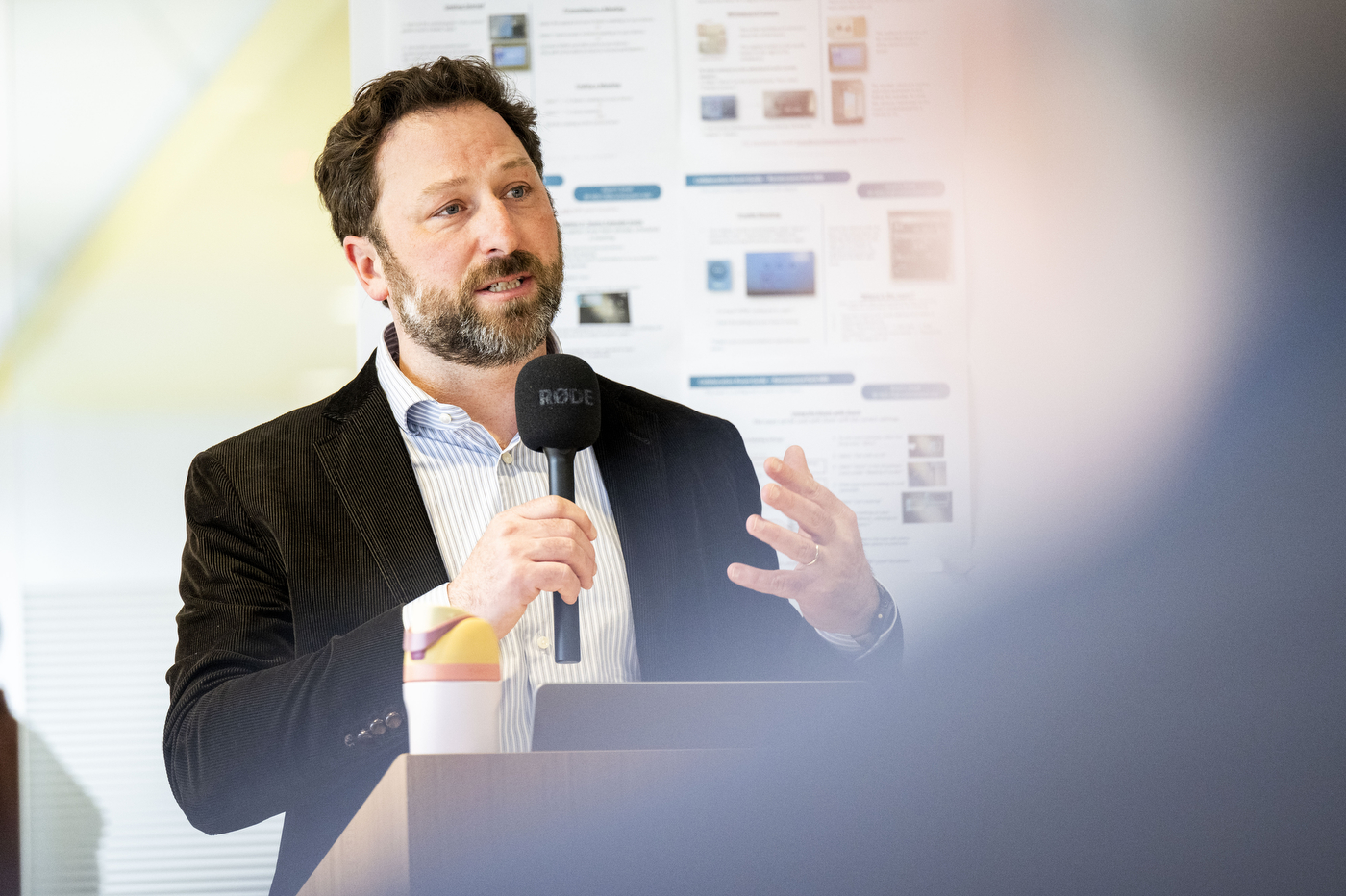 A bearded man in a dark blazer speaks into a microphone, gesturing with his free hand.