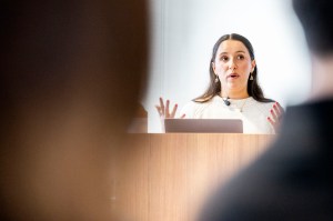 Melina Coy speaks at a podium during a presentation, gesturing with both hands while addressing an audience.