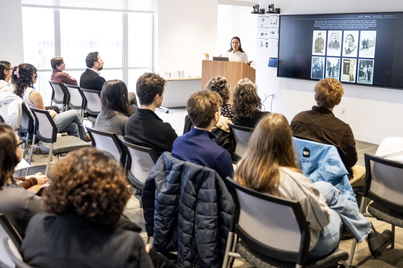 A wide shot of a modern conference room willed with attendees seated in rows, facing a presenter at the front near a screen.