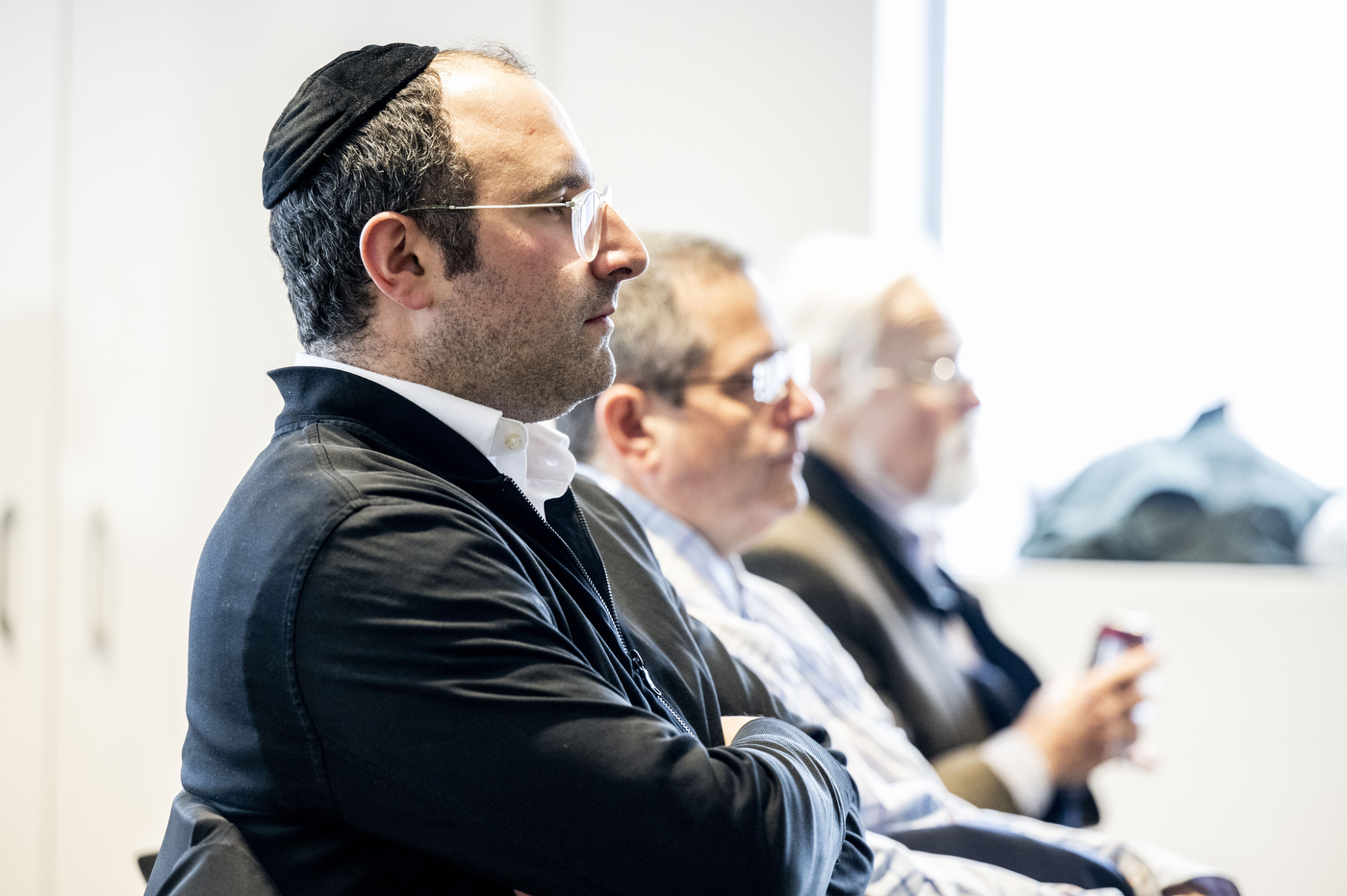 Three men seated in profile, listening to a presentation. The man in the foreground wears a dark suit and a kippah.