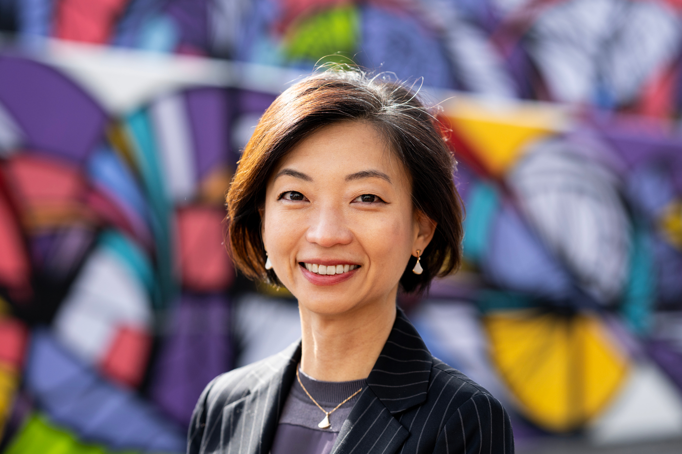 Portrait of Sangah Bae standing outside in front of a blurred wall painted with a colorful, geometric mural. 