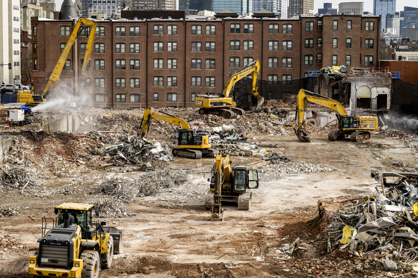 A wide view of the construction site showing multiple vehicles taking down the final pieces of the former Matthews Arena.