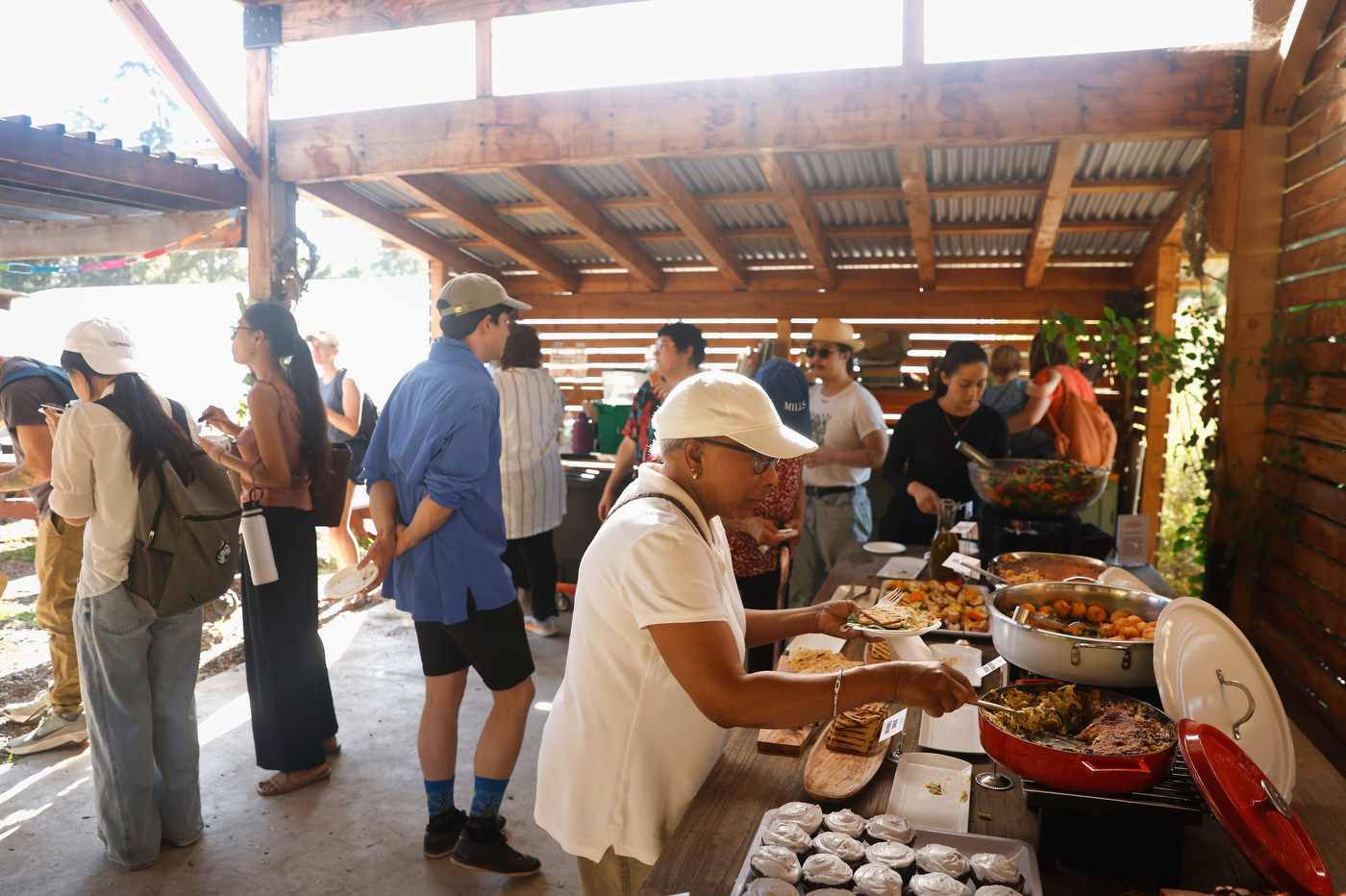 A group of people serve and receive food at outdoor tables under a covered pavilion on a sunny day. 