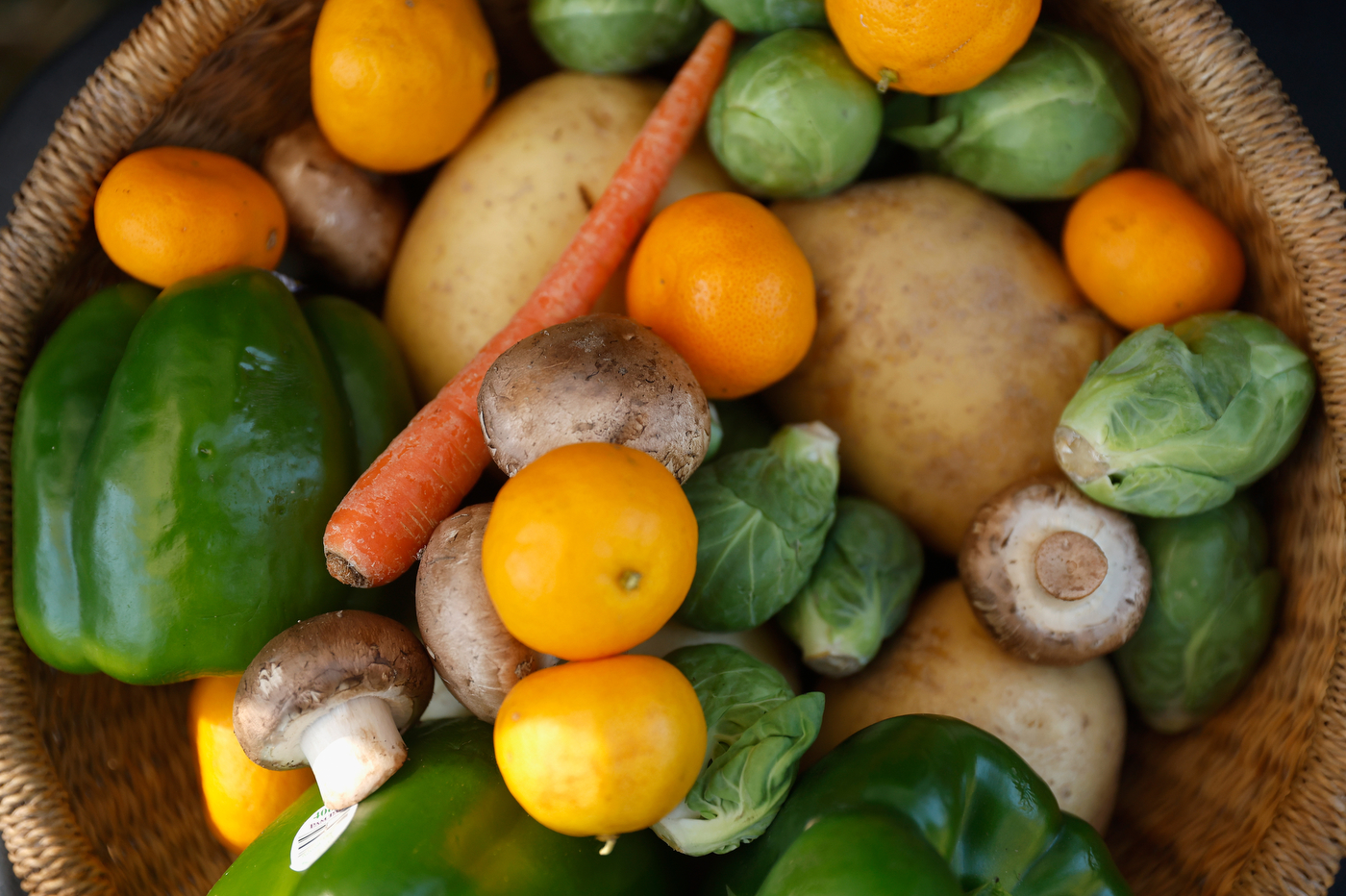 A close-up overhead of a basket filled with fresh vegetables and fruit, including lemons, carrots, potatoes, bell peppers, mushrooms, and Brussels sprouts. 