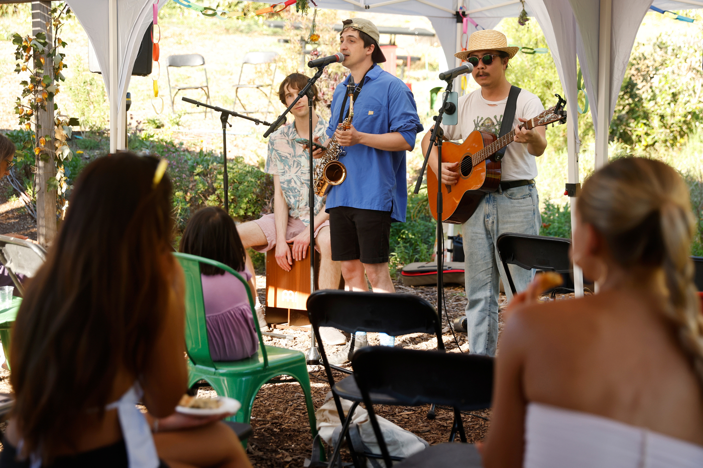 Three people play musical instruments, including a guitar and a saxophone. They stand in front of microphones. People in the audience sit in chairs. 