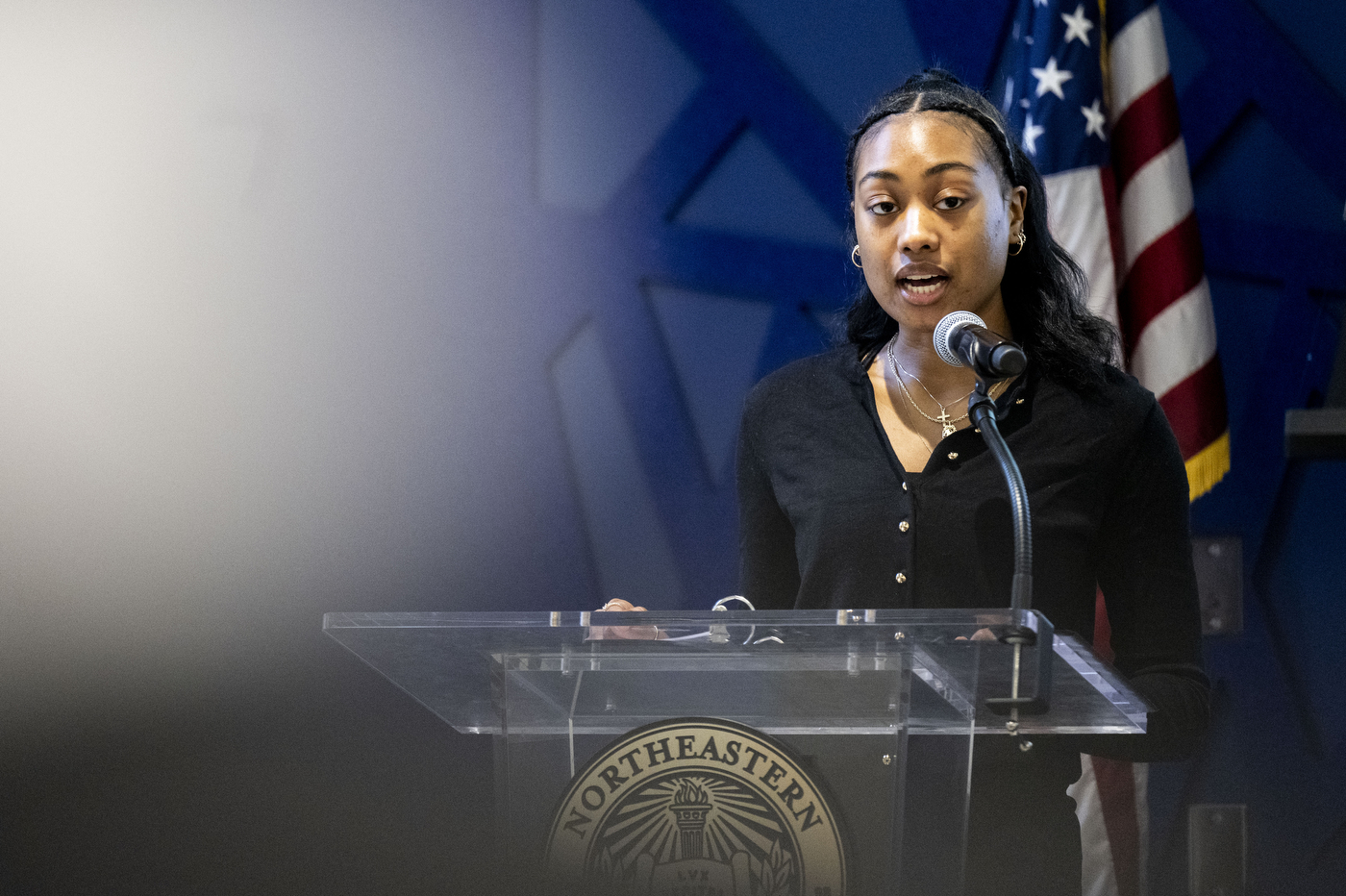 A woman speaks into a microphone at the Wellness & Wisdom from Women Who Build NU Alumni Table Talk event at the John D. O'Bryant African-American Institute in Boston, with an American flag visible behind her.