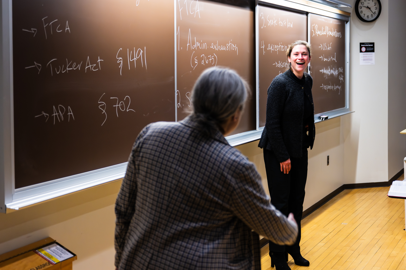 A person standing in front of a chalkboard smiles at another person.
