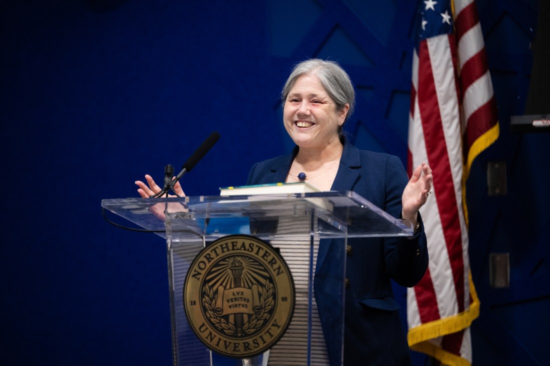 Beth Winkelstein gestures with her hands and smiles while speaking at a podium with an American flag visible behind her. 