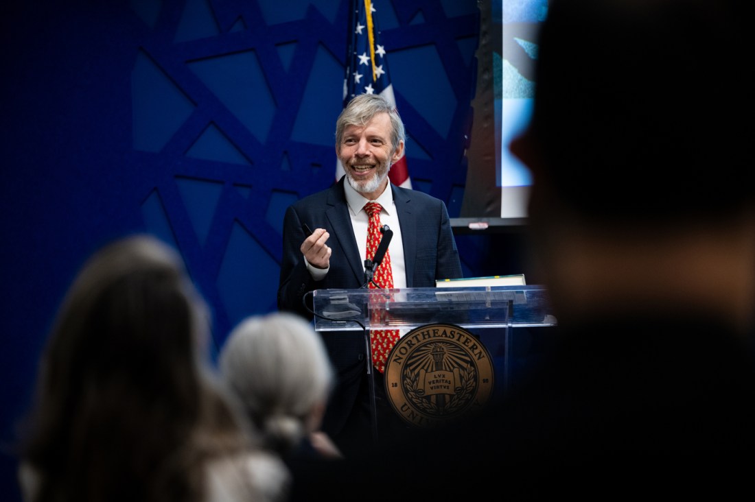 Koen Pauwels wearing a suit and red patterned tie smiles and gestures while speaking at a podium, with an American flag standing behind him. 
