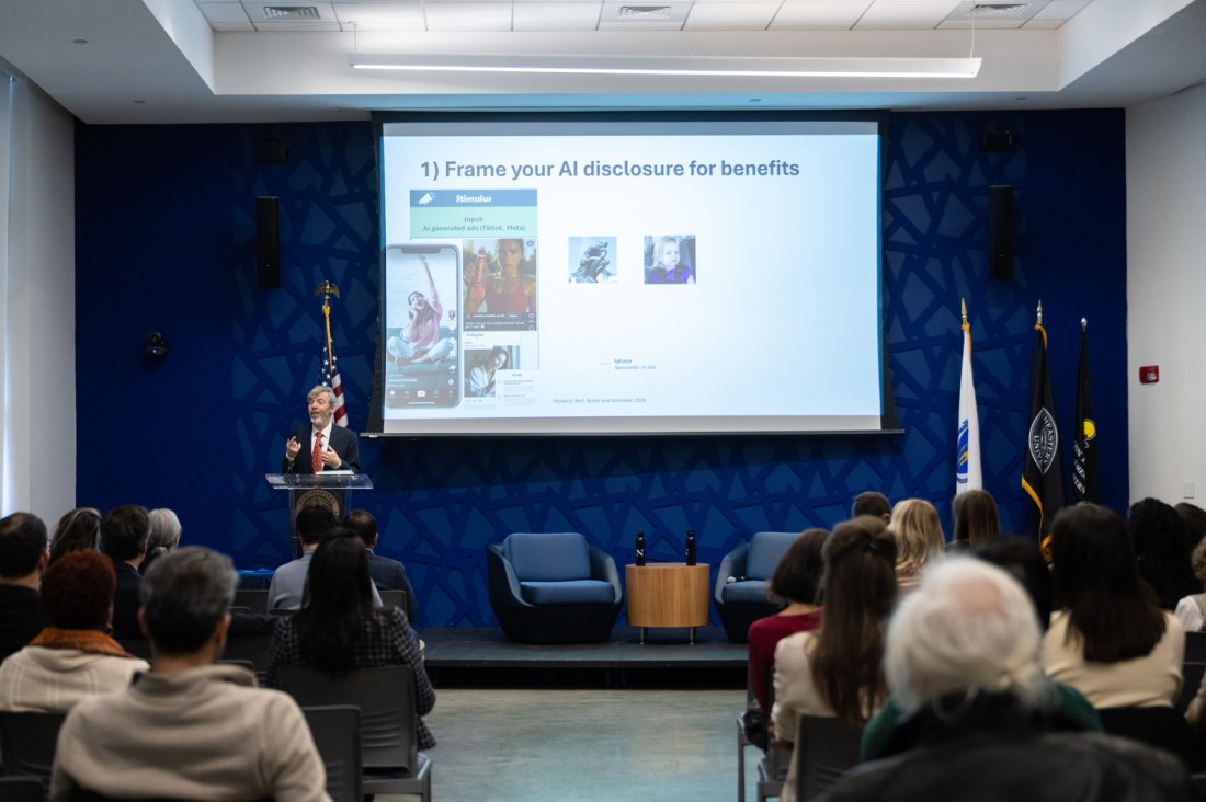 A wide shot of an auditorium-style room where an audience faces Koen Pauwels standing at a podium and a large projection screen behind him with a slide that says 'Frame your AI disclosure for benefits'. 