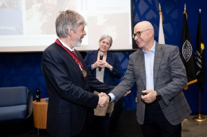 Koen Pauwels and President Aoun shake hands on stage in front of a presentation screen, while a third person standing behind them holds a microphone and looks on. Koen Pauwels is wearing a red ribbon with a medallion around his neck.