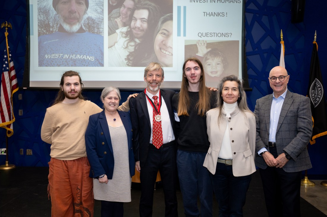 Koen Pauwels wearing a dark blazer, red tie, and Klein Lecturer medallion poses with a group of five other people, including Beth Winkelstein and President Aoun, in front of a presentation screen displaying several images and text that says 'Invest in Humans. Thanks! Questions?' at a Northeastern event. 