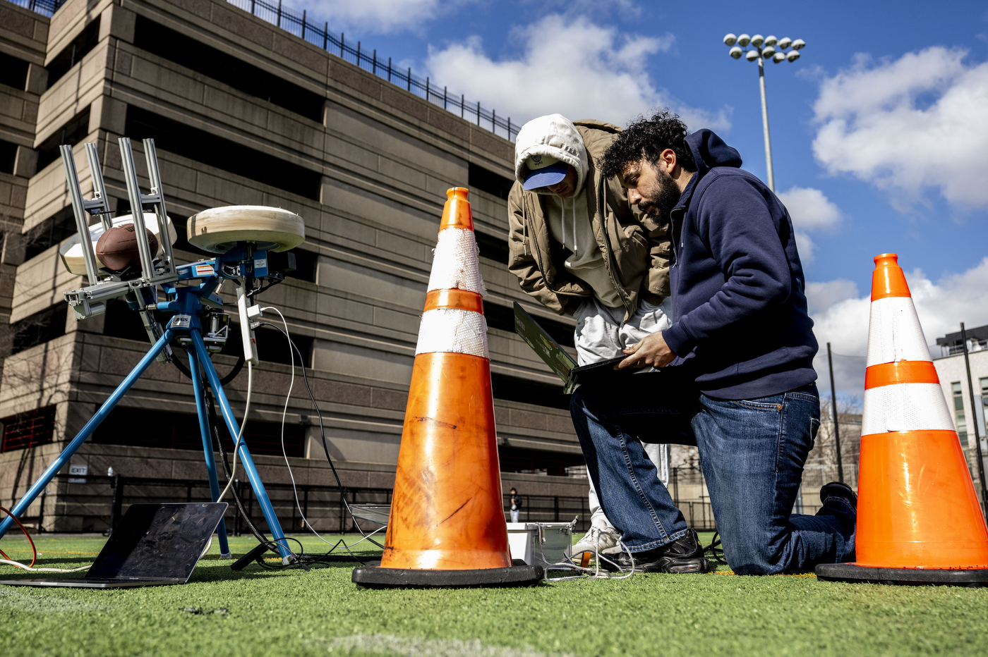 Two students set up an autonomous football-launching machine outside on a turf field. One is kneeling with a laptop resting on his knee, while the other looks over his shoulder. Traffic cones are in the foreground of the photo. 