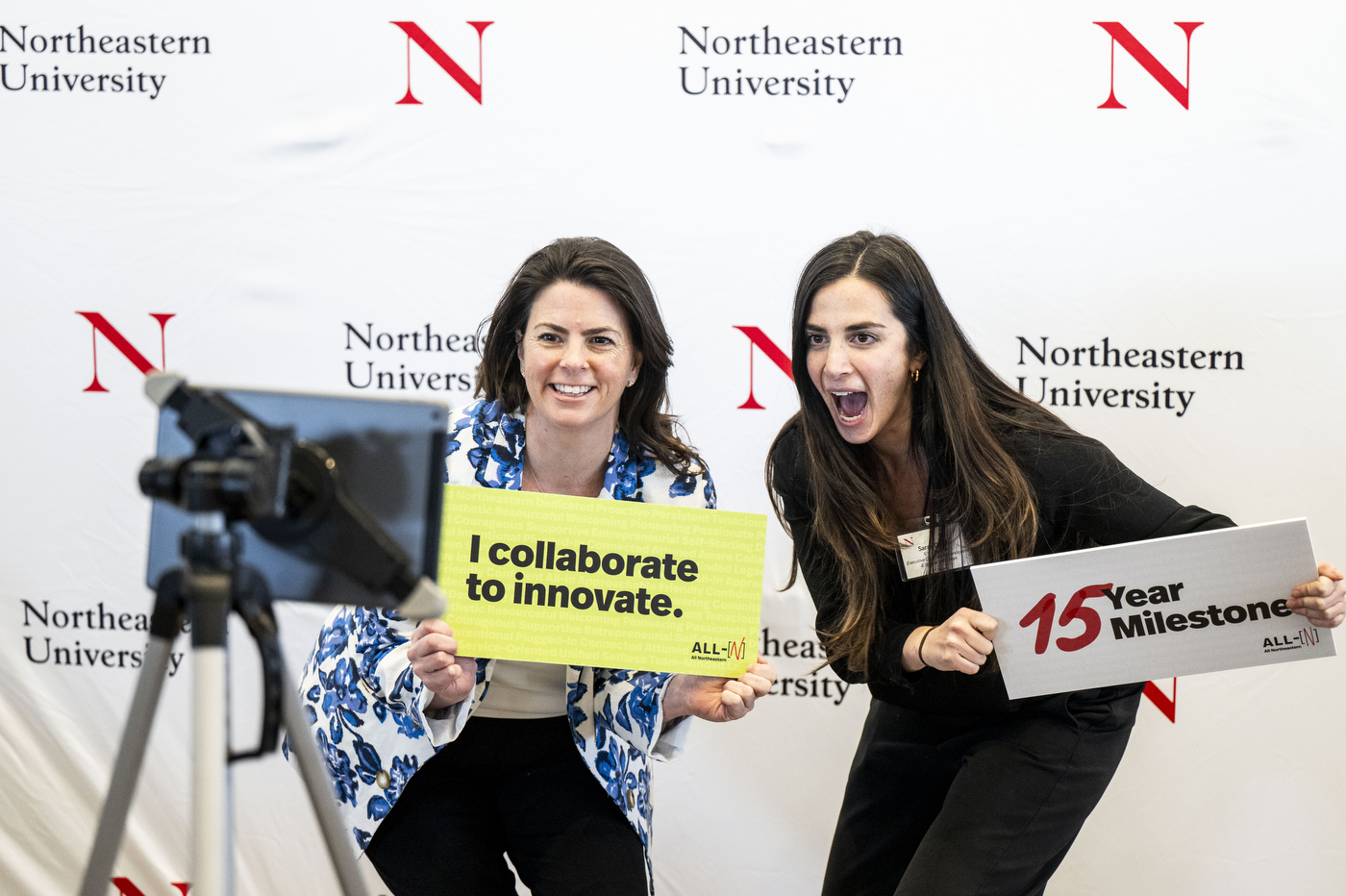 Two people hold up signs and pose for a photo in front of a backdrop that says "Northeastern University" and has the "Northeastern N" on it. 