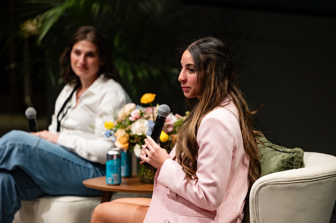 A woman in a pink blazer speaks into a microphone during a panel discussion, with another panelist seated beside her.