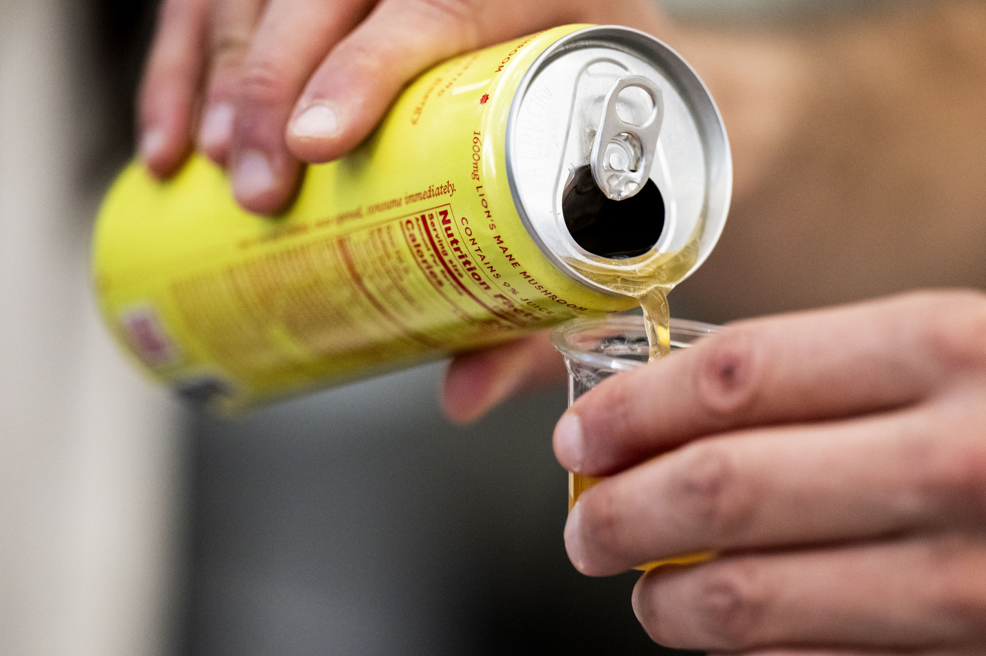 Close up of a person's hands pouring a yellow tinted energy drink from a tall, slim, yellow can into a small plastic cup. 