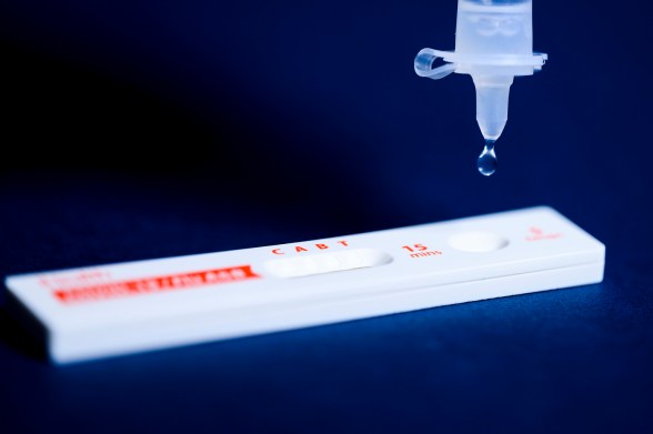 A COVID-19 test lying flat with a droplet falling from a syringe.