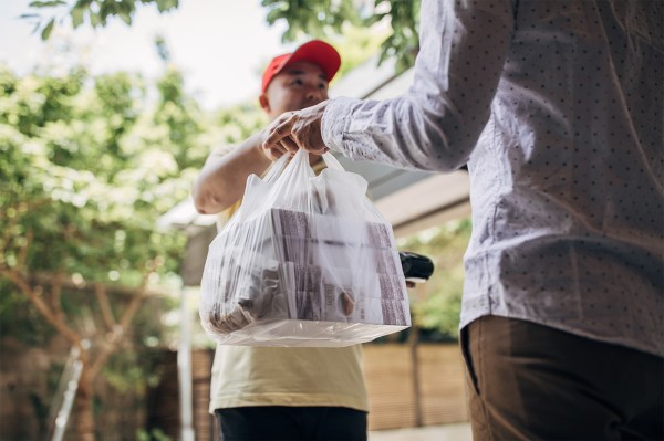 A delivery driver wearing a red baseball cap hands a plastic bag full of food to a customer. They are standing outside.