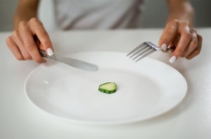 A person holds a knife and fork above a single slice of cucumber on an otherwise empty plate.