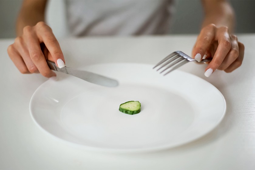 A person holds a knife and fork above a single slice of cucumber on an otherwise empty plate.