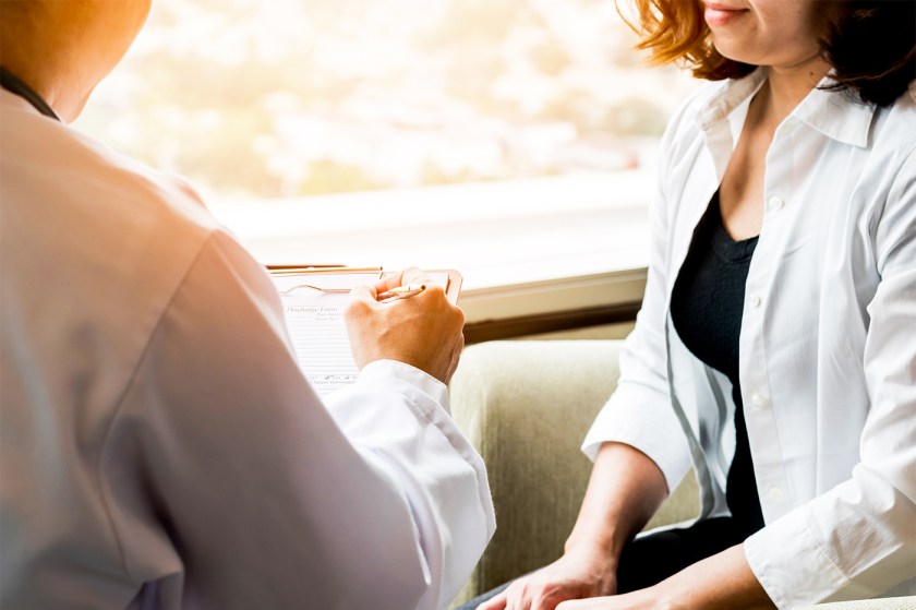 A physician in a white lab coat takes a note while a person in a white shirt sits next to them.
