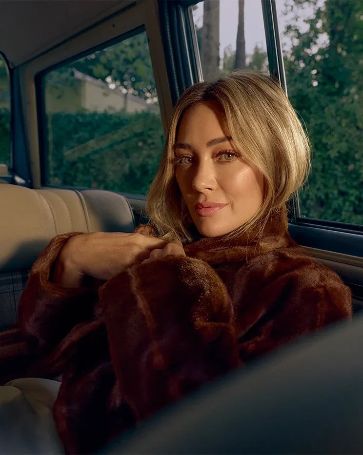 Hilary Duff poses in the backseat of a car, leaning her head against the window as the sun streaks across her face.