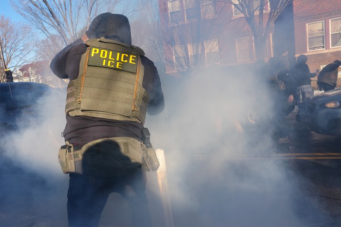 An immigration officer wearing a vest that says 'POLICE' on it in yellow on the back stands in a cloud of tear gas outside in Minneapolis.