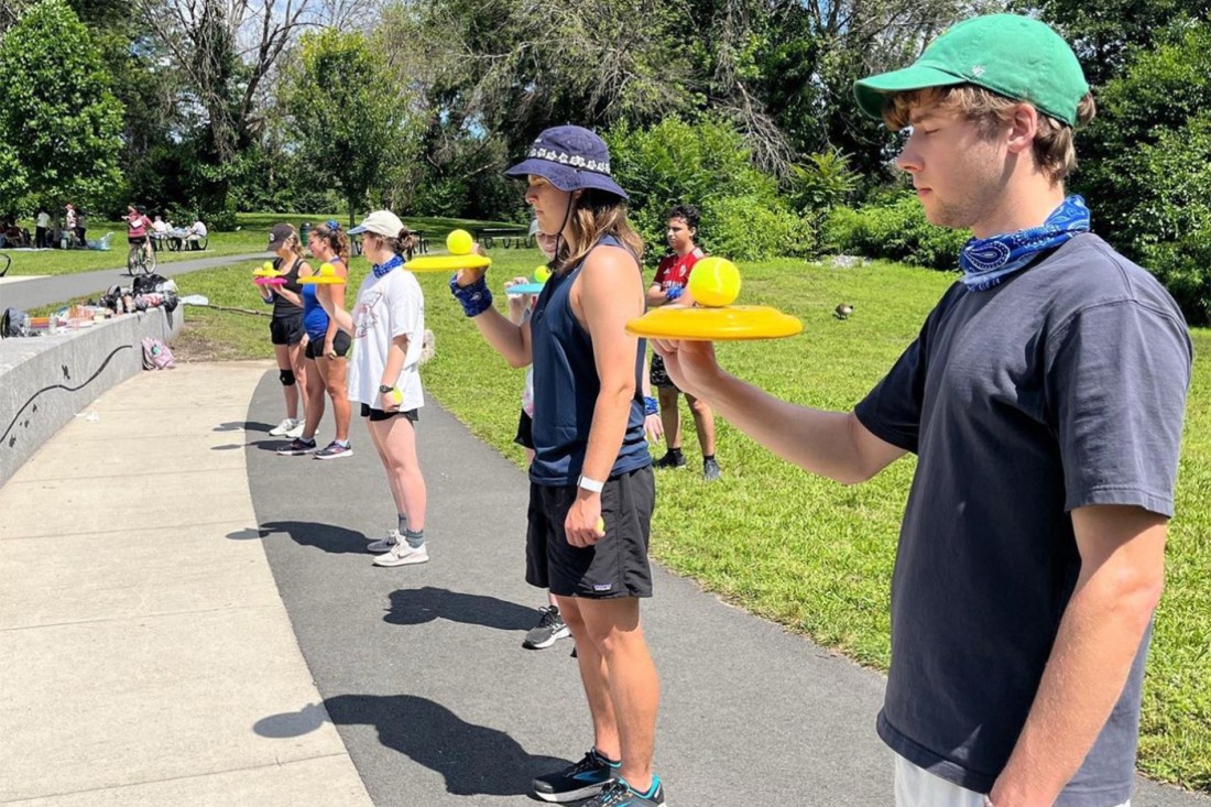 A line of four Survivor Northeastern contestants stand next to one another while trying to balance a ball on a frisbee.