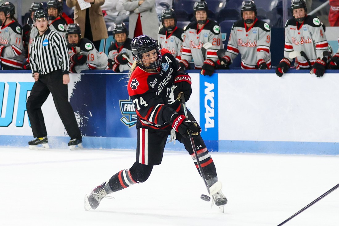 Northeastern skater controls the puck along the boards with teammates on the bench behind.
