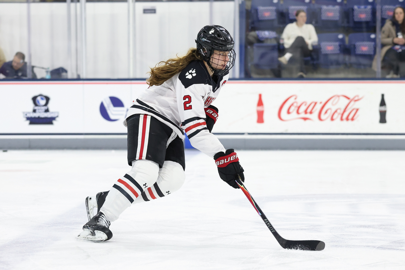 A hockey player in a white jersey with black and red stripes skates forward while controlling the puck on the ice during a game.