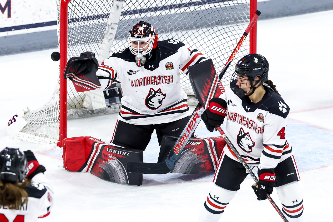 A goaltender in a white Northeastern jersey drops to their knees in front of the net to make a save as a teammate skates nearby during live play.