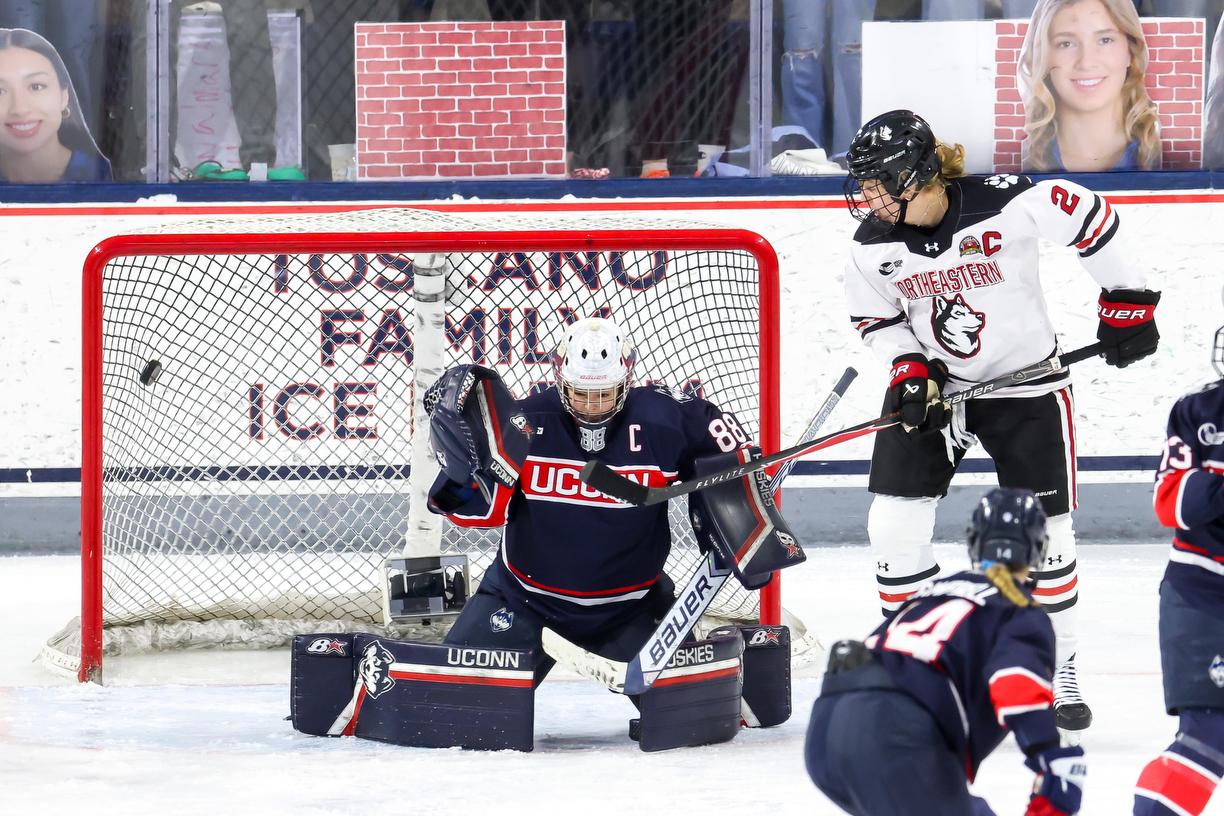 A UConn goaltender drops to their knees in front of the net to block a shot as a Northeastern player waits near the crease and another UConn skater moves into the play during a hockey game.