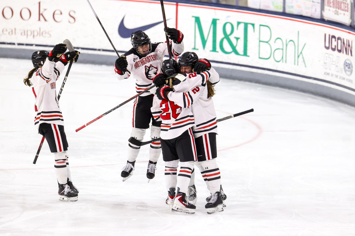 Four Northeastern hockey players celebrate on the ice, raising their sticks and hugging after a goal during a game.