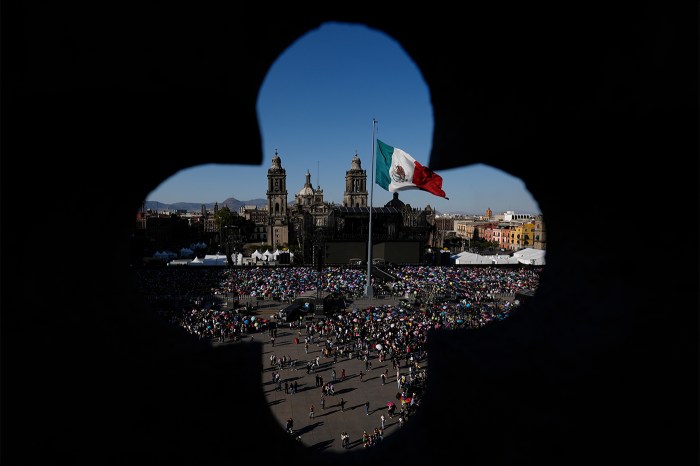 A photo taken from inside a building looking through a clover-shaped hole, darkening the surrounding edges. Through the gap, many people gather in a square with the Mexican flag flying above them.