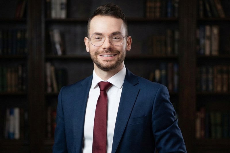 Ryan Rad, in a suit and tie, poses for a portrait in front of a bookcase.