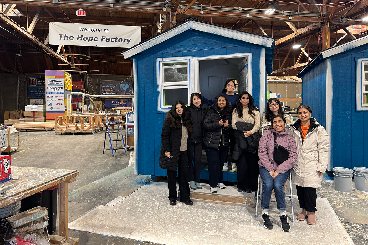 Students from Agents of Social Impact pose in front of a tiny home shelter in a warehouse. In the background a banner hangs that says 'Welcome to The Hope Factory.'