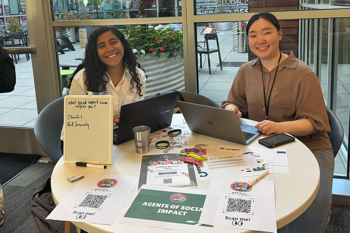 Two students sit at a table tabling for Agents of Social Impact, with flyers and QR codes displayed in front of them.