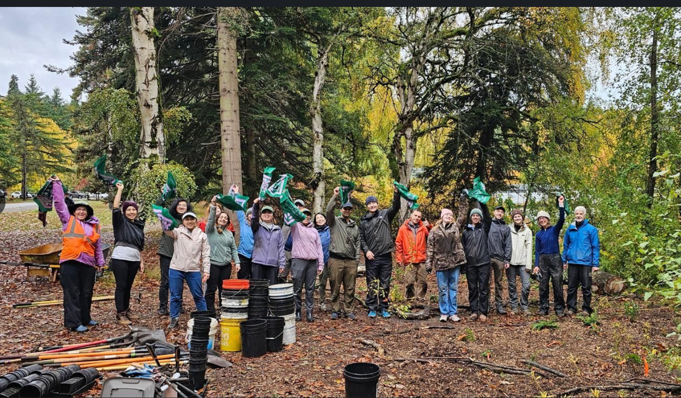 A large group of students and community voulnteers raised their hands in a wooded area during an outdoor volunteer event.