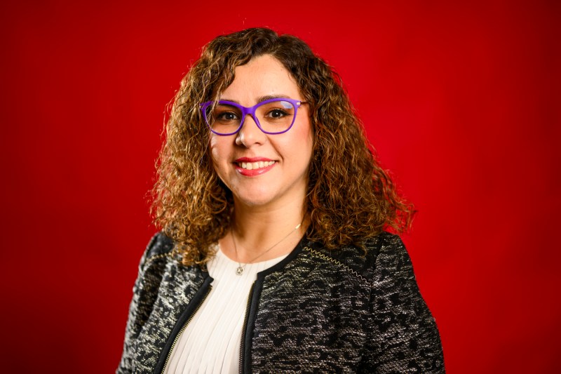 Serena Alexander, with long hair and purple glasses, stands against a red background.