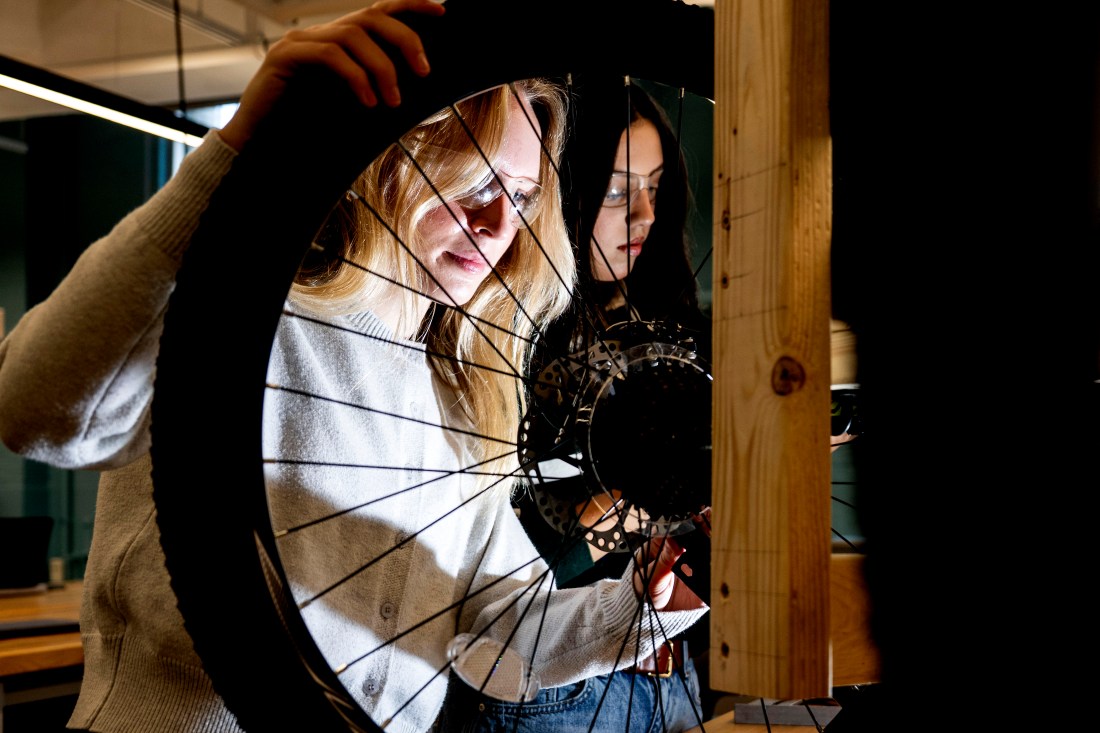 Two students wearing safety glasses inspect the hub and spokes of a bicycle wheel next to a wooden frame component of an affordable wheelchair. 