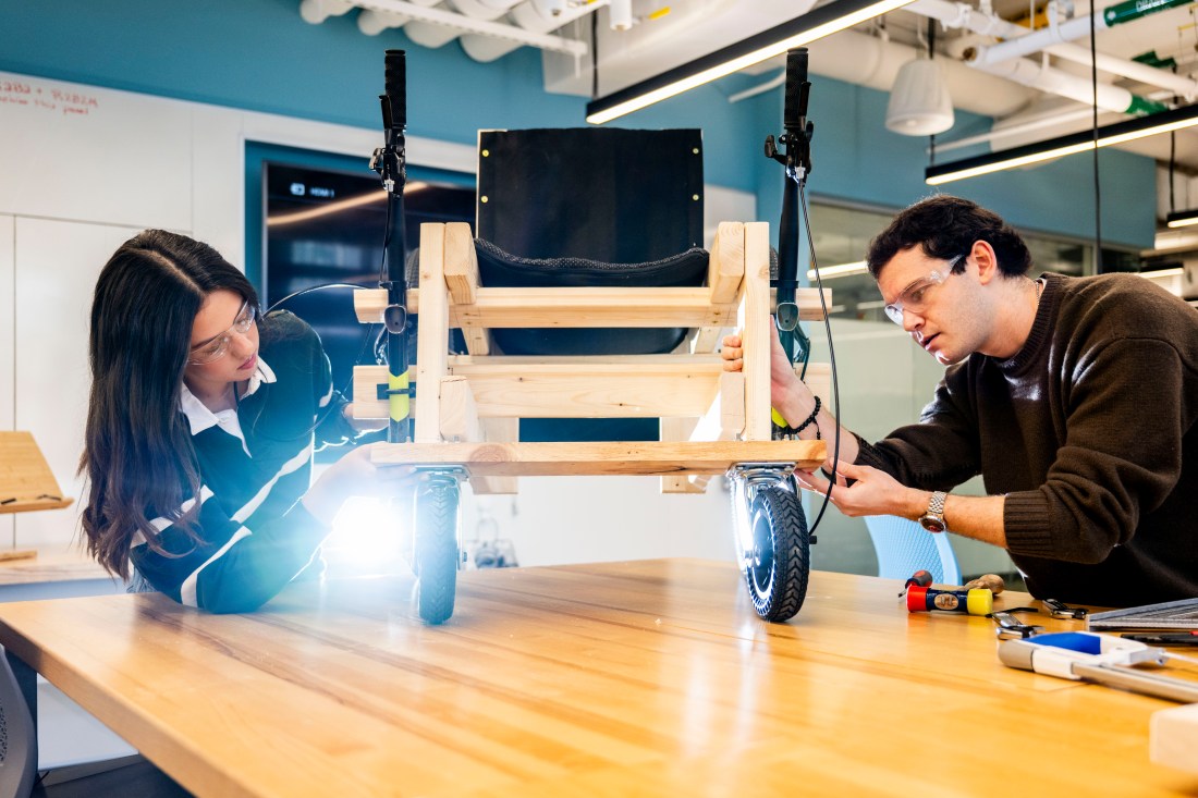 Two bioengineering students examine a wooden wheelchair prototype with bicycle wheels on a workbench. 