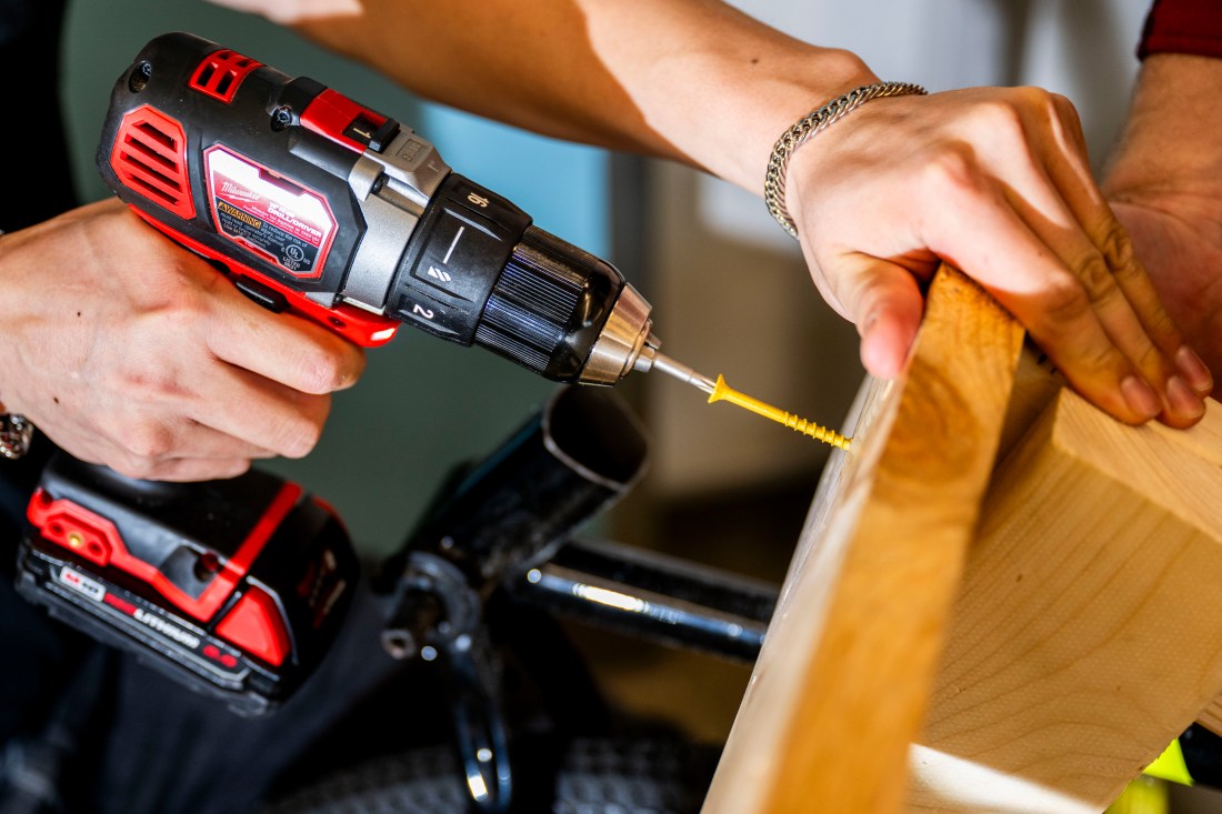 A close-up of a students hands using a power drill to screw in a wooden component of a wheelchair frame. 