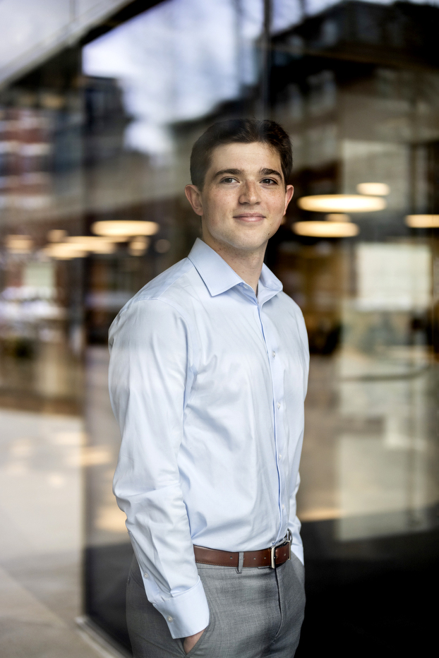 Portrait of Ethan Bengelsforf in a light blue button-down shirt smiling, photographed in a modern interior space with blurred ceiling lights in the background.