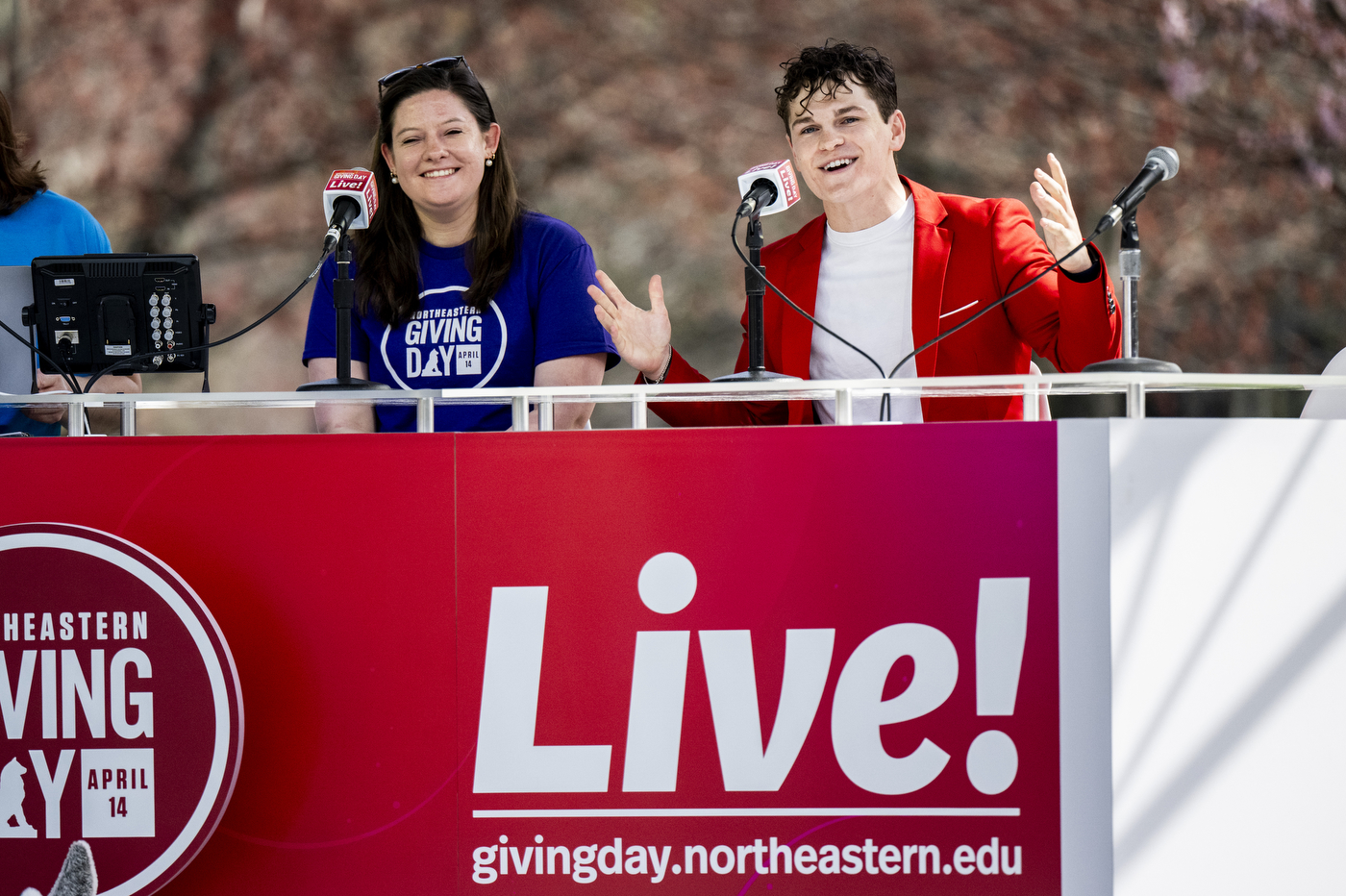 Two hosts sit at a Giving Day live broadcast desk outdoors. One is wearing a red blazer and gesturing while speaking into a microwave. 