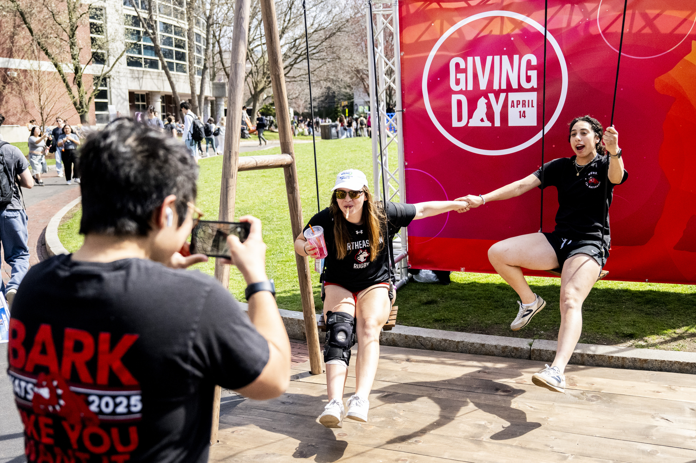 Two students pose on a swing set in front of a Giving Day banner while a third person takes their photograph. 