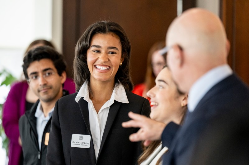 A smiling student stands beside President Aoun at the Presidential Ambassador Senior Reception, with another student visible in the background.