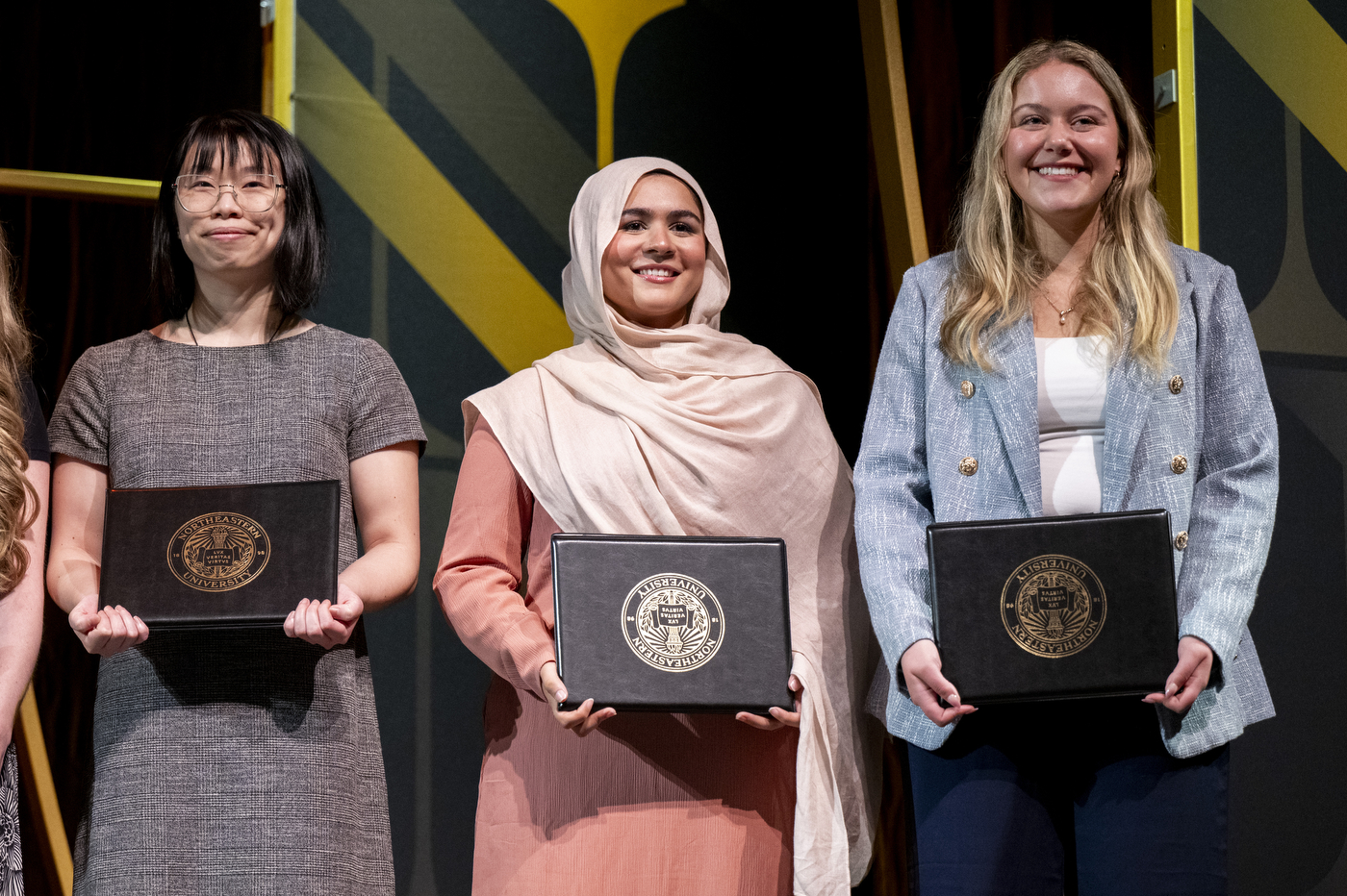 Three Northeastern University students smile and hold award portfolios with the university seal at the Academic Honors Convocation ceremony.