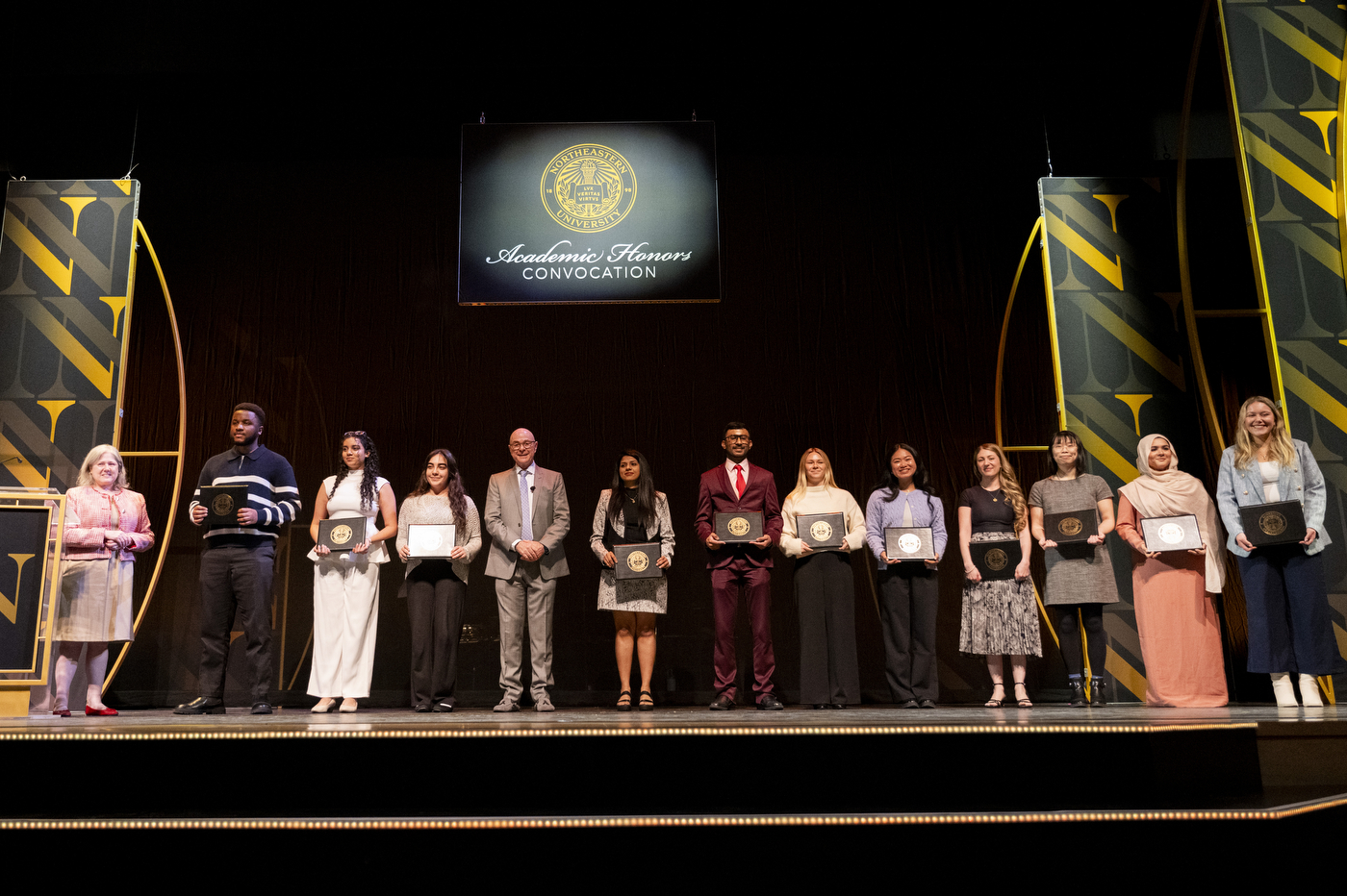 A group of Northeastern University honorees stands in a line on stage at the Academic Honors Convocation, with the university seal projected on a screen behind them.